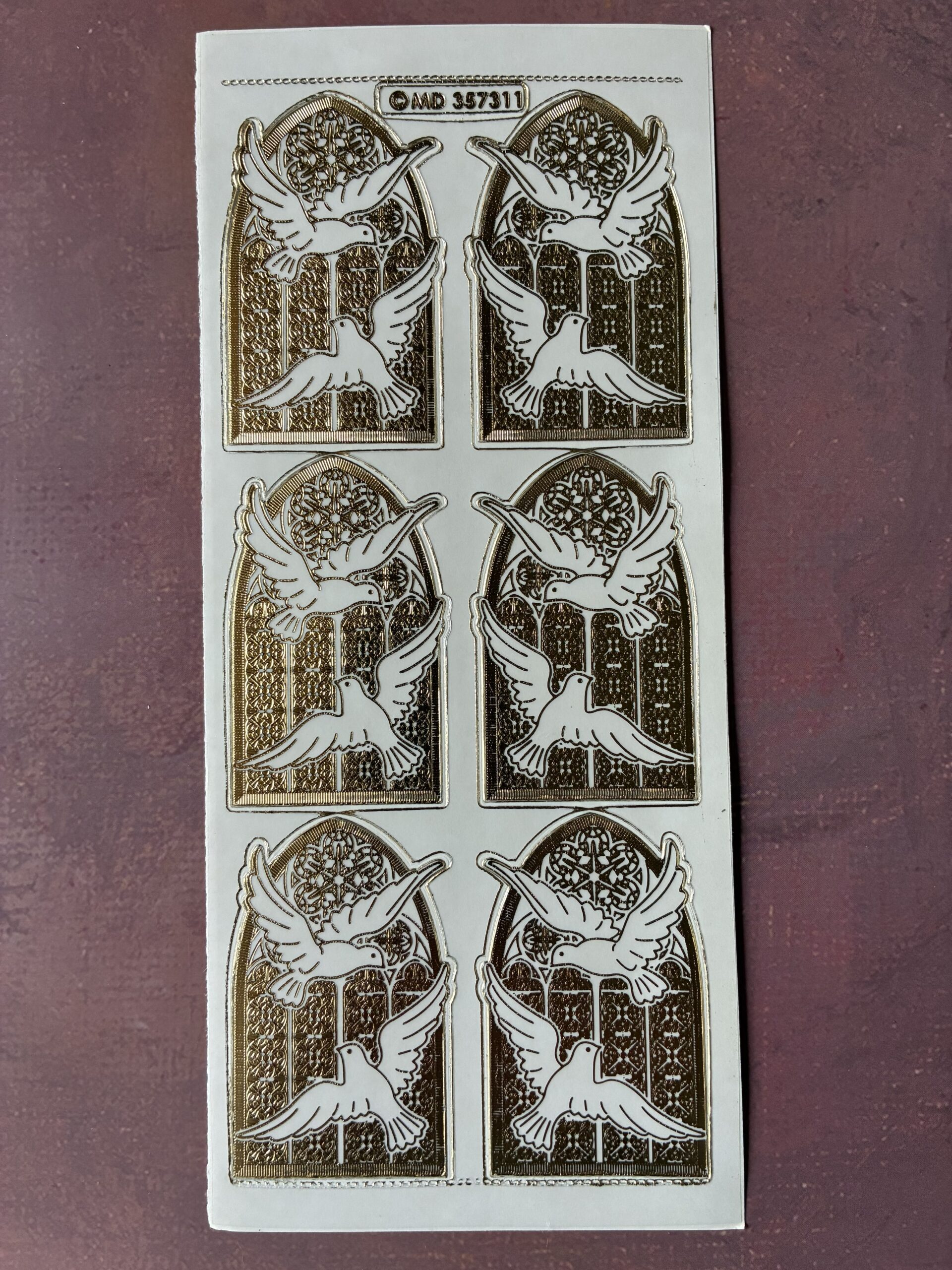 Doves Window Transparent Gold. Religious themed peel off stickers with 6 images of a pair of doves in front of a stained glass window in a gold colour.